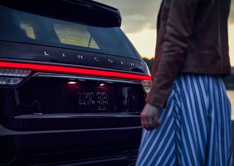 A person is shown near the rear of a 2024 Lincoln Aviator® SUV as the Lincoln Embrace illuminates the rear lights | Northgate Lincoln in Port Huron MI