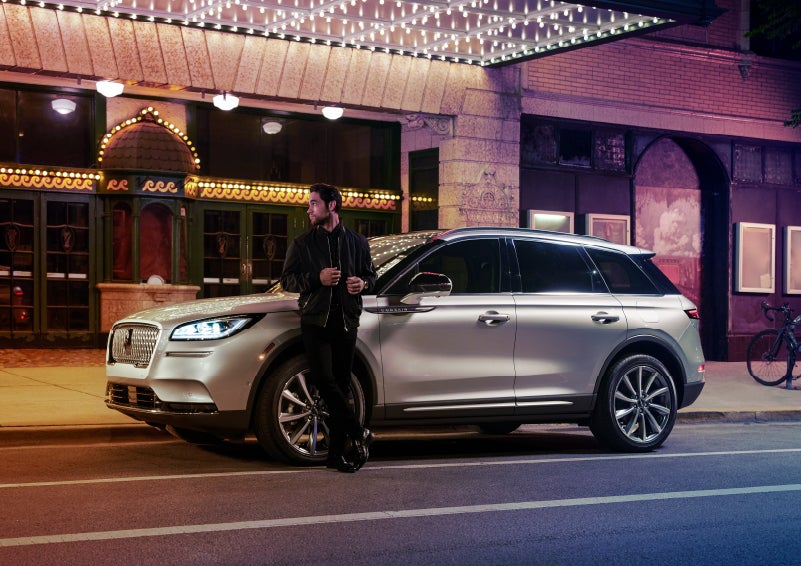 A 2022 Lincoln Corsair SUV is parked outside a theater as the driver relaxes against the frame and lights illuminate the floating roofline and body | Northgate Lincoln in Port Huron MI