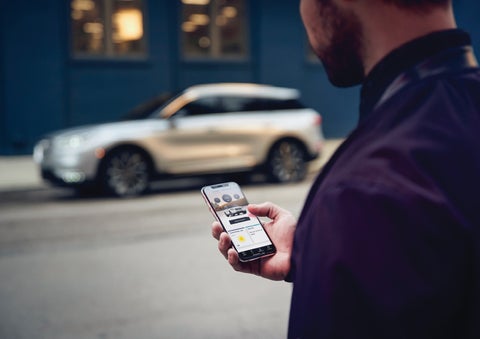 A man is holding his phone looking at the Lincoln Way™ App and we see his 2022 Lincoln Corsair SUV in the background against a blue brick wall | Northgate Lincoln in Port Huron MI