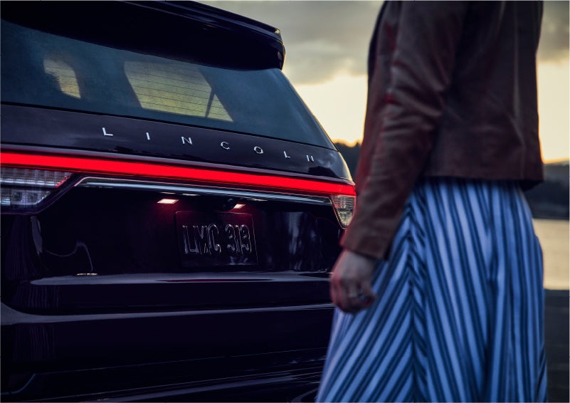 A person is shown near the rear of a 2023 Lincoln Aviator® SUV as the Lincoln Embrace illuminates the rear lights | Northgate Lincoln in Port Huron MI
